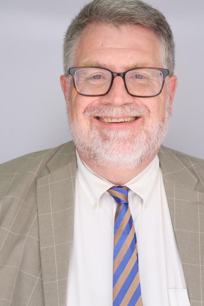 Headshot of Steven Brady against a gray background. He is wearing a tan suit with a buff and blue tie.